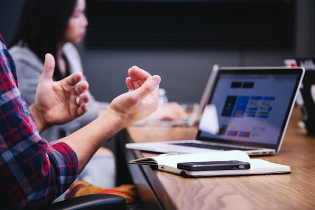 Consultant gesturing while discussing strategy at a table with an open laptop and notebook