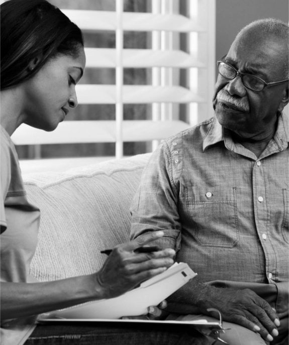 Black-and-white photograph of a caregiver writing on a clipboard beside an older man seated on a sofa